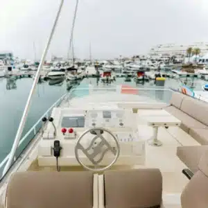 The helm of a boat with a steering wheel and navigation instruments, set against a marina backdrop filled with moored boats on an overcast day.