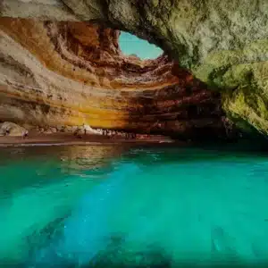 A stunning coastal cave with sunlight highlighting layered rock formations, turquoise water, and people enjoying the sandy beach inside.