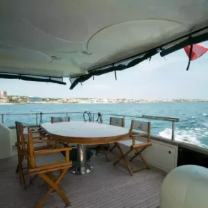 Outdoor dining setup on yacht deck with ocean view, wooden chairs, and safety railings, near coastline under clear sky.