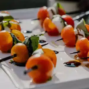 A creative arrangement of gourmet appetizers with orange fruit or vegetables, leafy green garnish, and dark sauce on skewers at a high-end dining event.