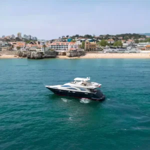 A luxurious motor yacht cruising near a coastline with a sandy beach and coastal town under clear and sunny weather.