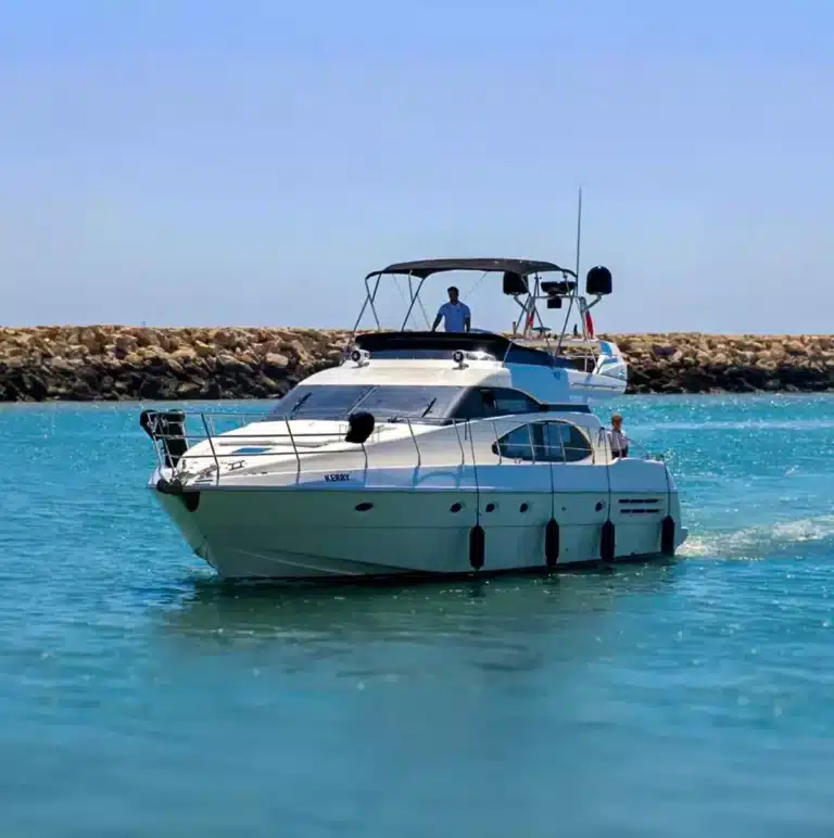 Luxurious motor yacht named Kerry on clear blue water with people enjoying a sunny day, near a breakwater made of large boulders.