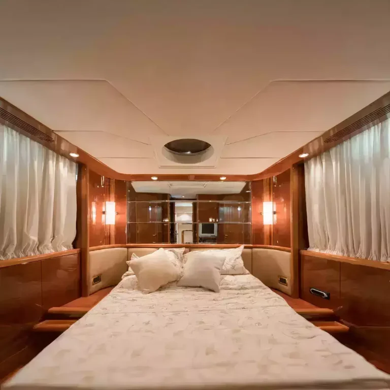 Luxurious yacht cabin with polished wood cabinetry, white bedding, multiple pillows, and natural light through sheer curtains.