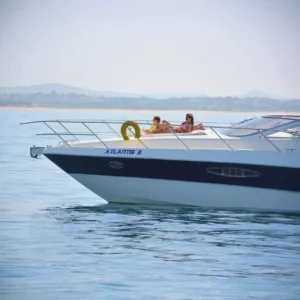 A luxury powerboat named ATLANTIS II on calm waters with two individuals lounging on the deck, near a hazy coastline.