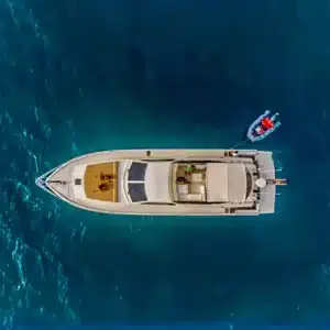 A bird's-eye view of a large yacht on varying shades of blue water, with a smaller dinghy tied to it and a few people aboard.