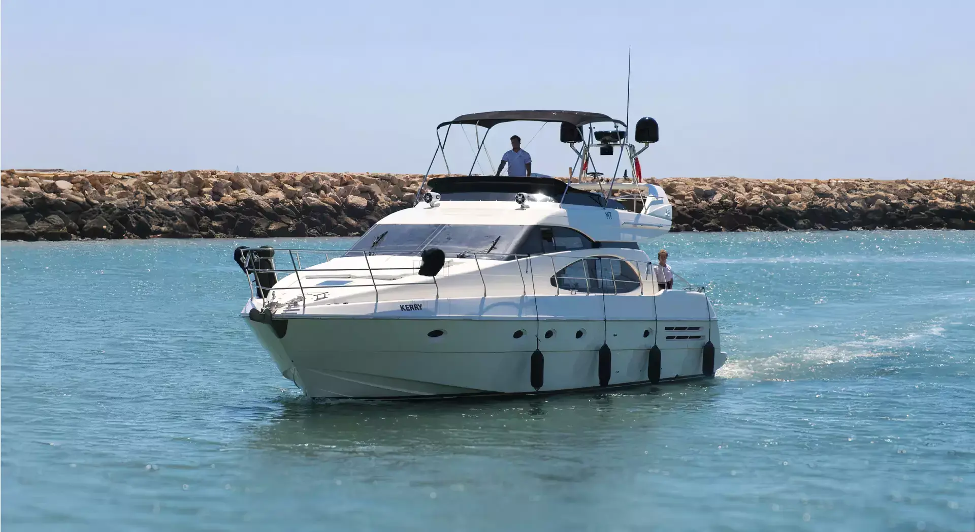 A white motor yacht with two people onboard cruising in calm blue water near a rocky breakwater under clear skies.