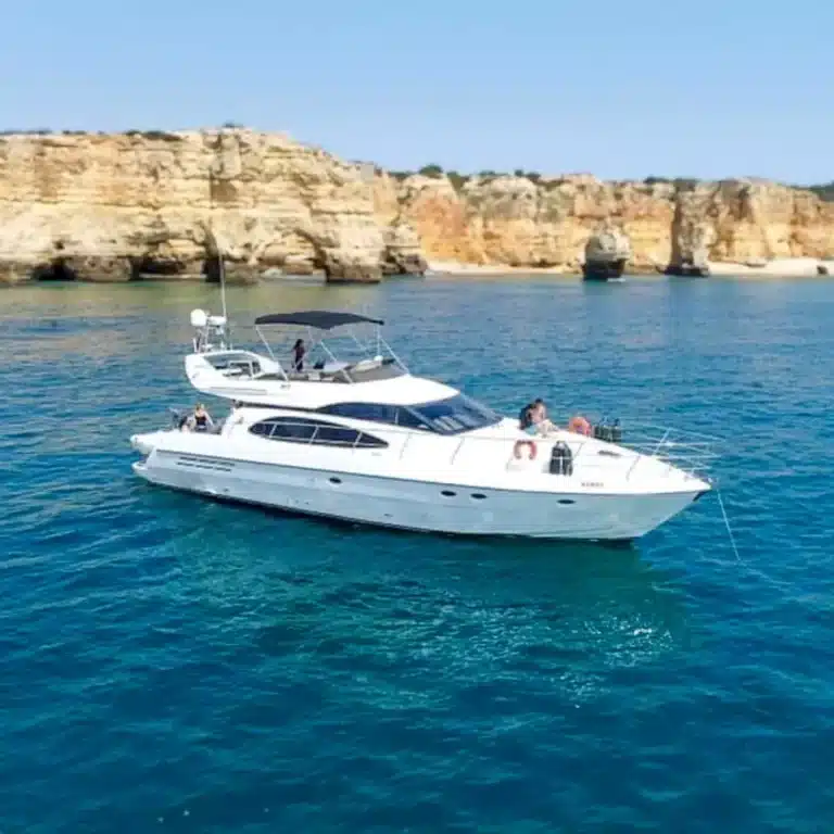 Luxury yacht floating on clear blue water with rocky coastline in background. Multiple people enjoying the sunny seaward excursion on the spacious yacht.