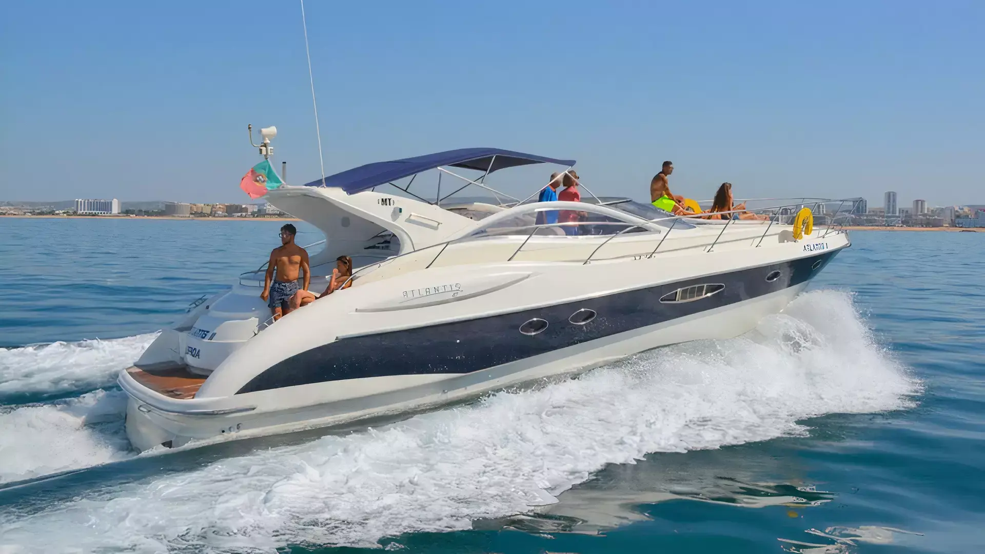 A white and navy blue motor yacht cruising through coastal waters on a sunny day, with people on board enjoying the view of the city skyline in the background.