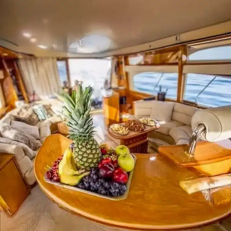 Interior of a luxury yacht showcasing a glossy wooden table with a fruit platter and snacks, cushioned seats, and ocean view.