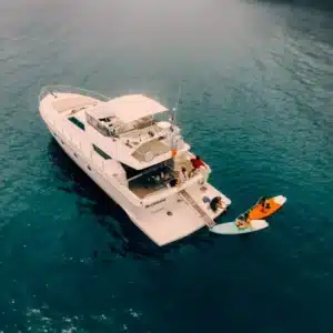 A large luxury yacht anchored in calm blue waters with people engaged in leisure activities like fishing and paddleboarding.