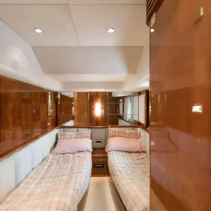 Interior of luxury yacht stateroom featuring two single beds with plaid bedding, high-gloss wood finish, recessed lights, and mirror for a spacious look.