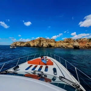 A person enjoying a serene moment on the bow of a cruising yacht with a rocky coastline and blue skies in the backdrop.