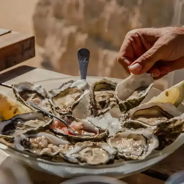 Plate of raw oysters on half shell served on ice, with a hand holding an oyster shell, lemon wedge, and small fork.