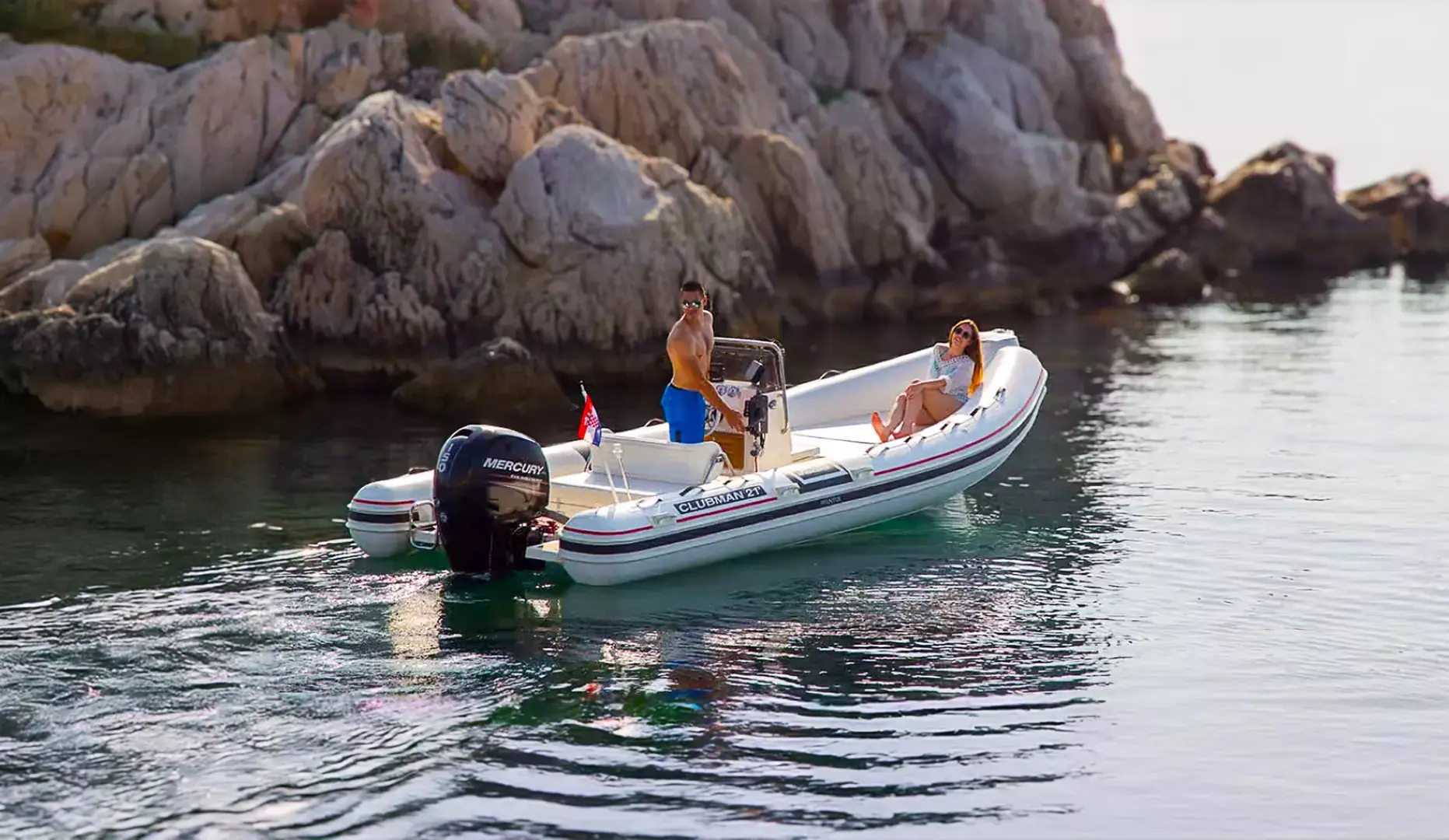 Two people enjoying leisure time on a Clubman 21 inflatable motorboat by a rocky shoreline in calm waters.