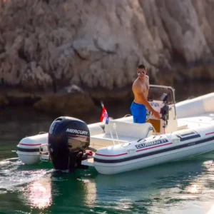 A shirtless man wearing sunglasses and blue swim trunks stands on a rigid inflatable boat with Mercury outboard motor near a rocky coastline.