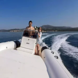 Two people enjoying a speedboat ride with a scenic coastline and clear sky in the background, creating a wake on the water.