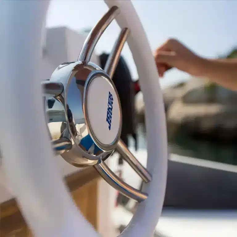 Close-up of a metallic and white ship's steering wheel with a blue and white logo, suggesting the act of piloting a yacht.