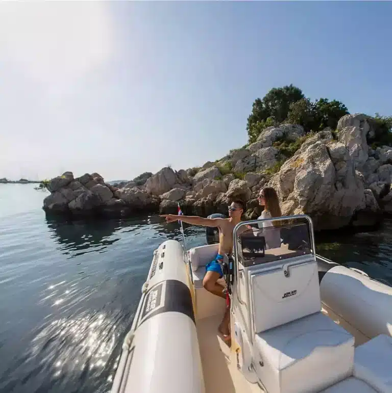 Two people enjoying a sunny day on a RIB boat, pointing towards rock formations in clear blue waters with other sails in the distance.