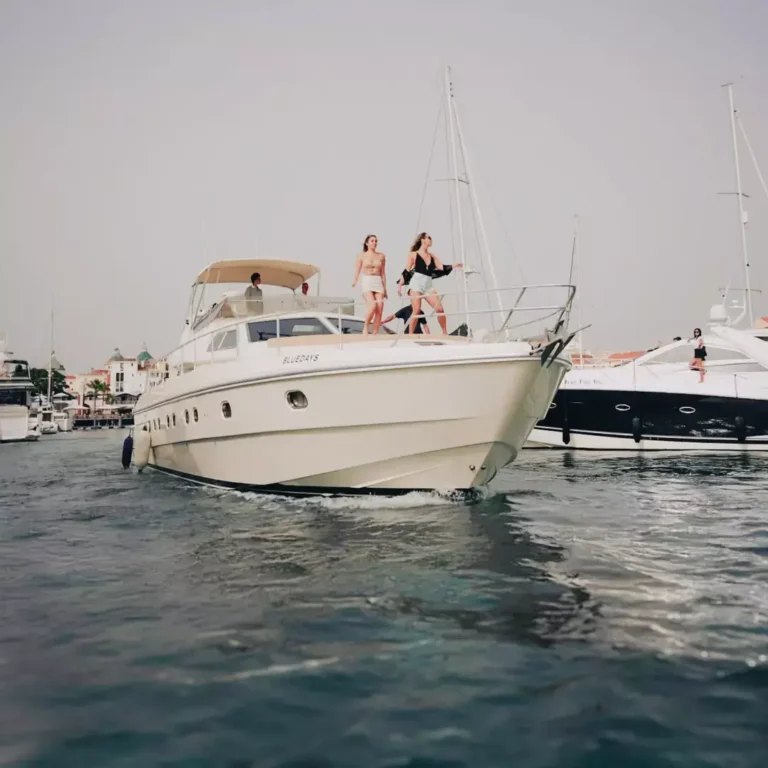 A white motor yacht with several individuals enjoying a leisurely day on the water, against an overcast sky and a backdrop of other boats.