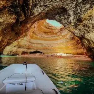 Yacht bow entering a sea cave with striated rock formations and sunlight streaming through the opening, illuminating the clear water and small beach.