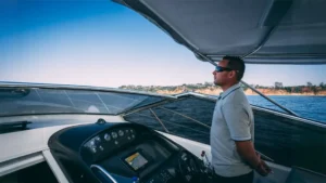 Person steering YOLO One yacht at Marina de Vilamoura under blue sky with coastline view.