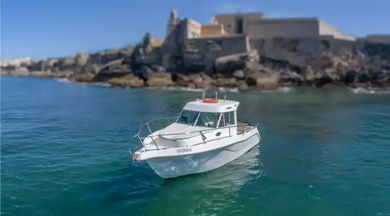 A small white motorboat named KATERINA B cruises through calm blue waters with a rocky coastline and historical architecture background.