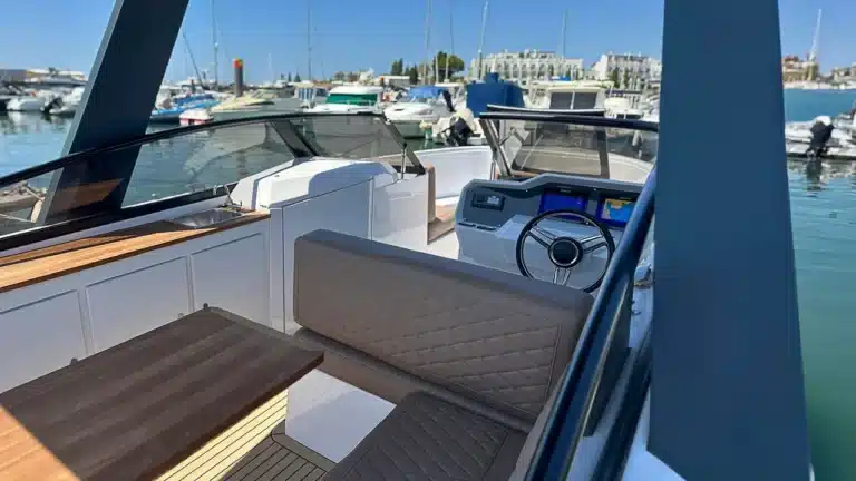 A modern yacht's helm showcasing a steering wheel, navigation display, quilted seat, and wood decking with boats in the marina and blue skies in the background.