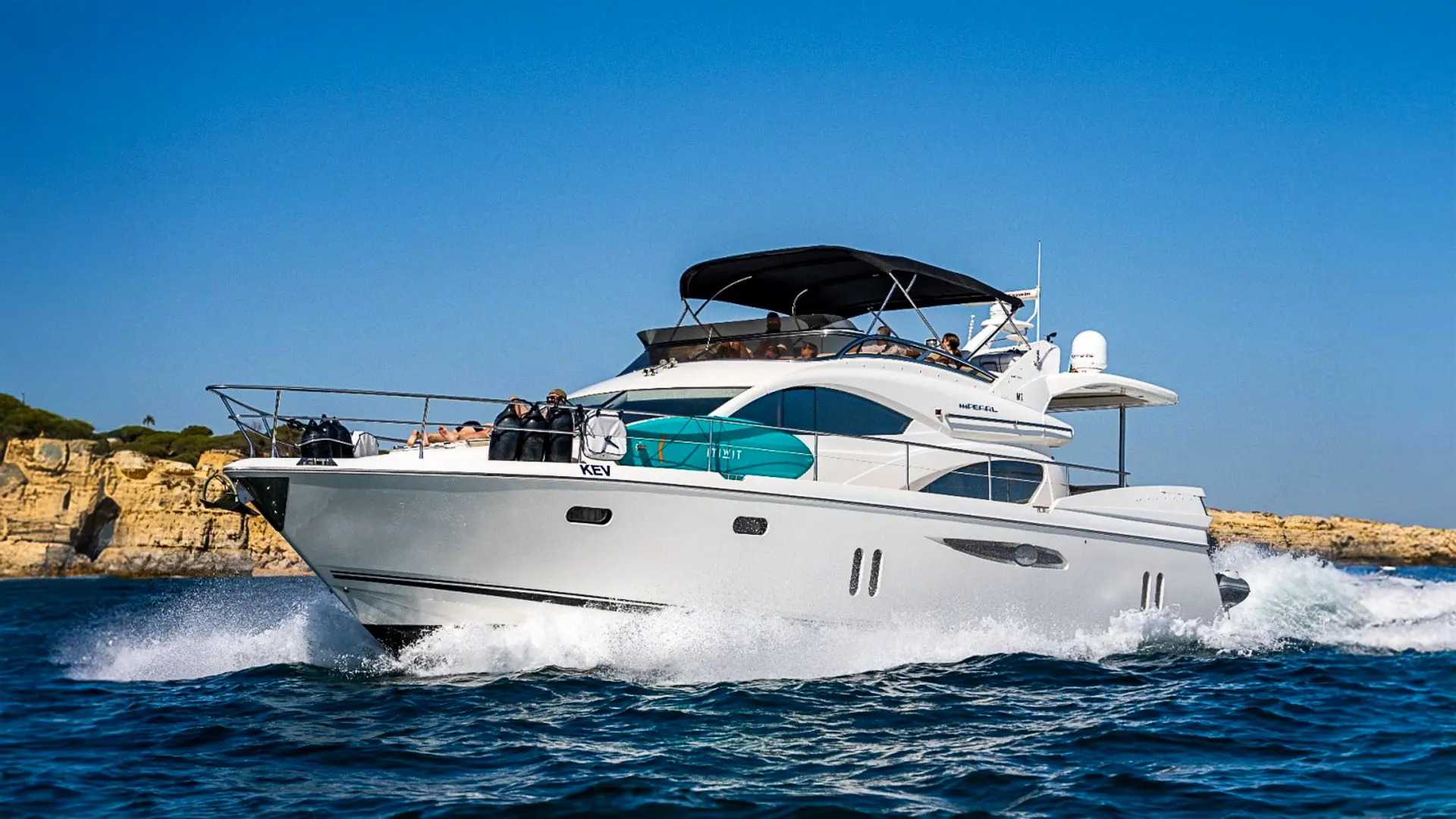 Luxury motor yacht KEV cruising near a coastline, showcasing its spacious design and amenities with people enjoying on the upper deck under a sunshade.