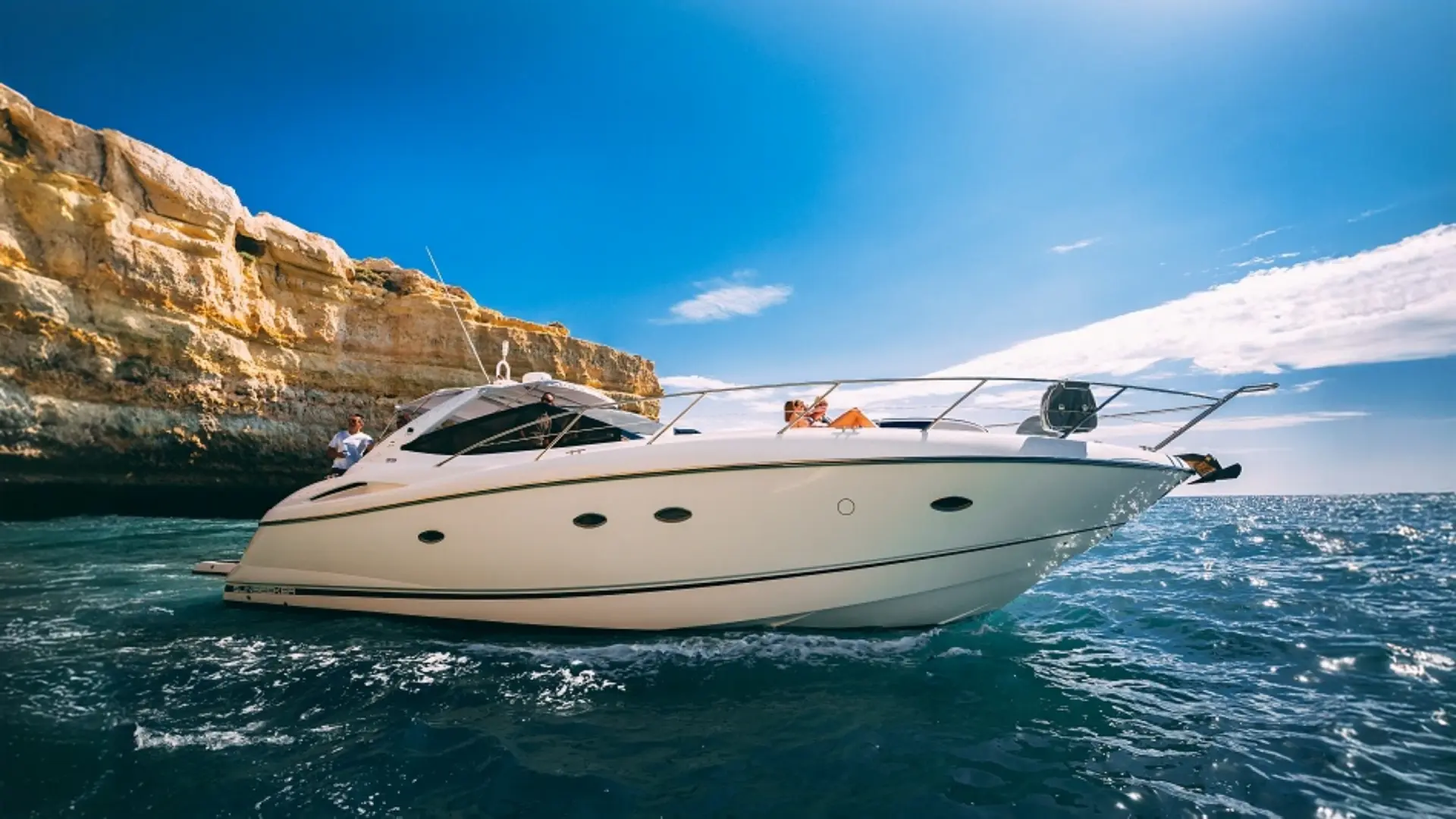 Luxury motor yacht YOLO One cruising near a rocky coastline under a clear blue sky with people enjoying themselves on a beautiful day.