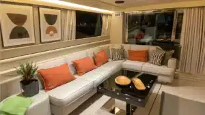 The living room of Katerina XL yacht in Cascais Marina at night, featuring a light-colored L-shaped sofa, abstract artworks, and warm ambient lighting.