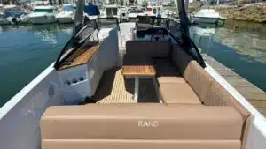 The deck of a modern motorboat with tan upholstery, teak-style flooring, and a wooden table, docked at a marina with calm water and clear sky.