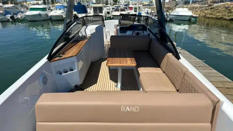 The deck of a modern motorboat with tan upholstery, teak-style flooring, and a wooden table, docked at a marina with calm water and clear sky.