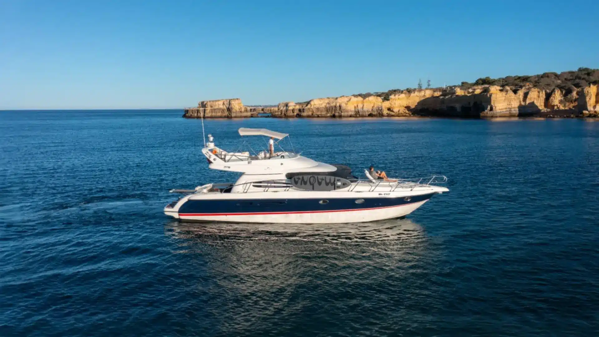 Mr Cliff, a motor yacht with white hull and blue stripe, cruising on calm blue seawater with a flag at the bow and a person sunbathing on the foredeck.