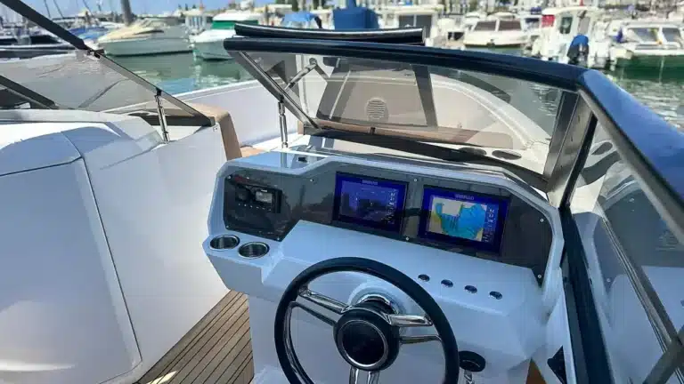 The helm station of a Rand Supreme 27 yacht featuring a modern control panel with navigation displays, wheel, and marina view on a calm day.