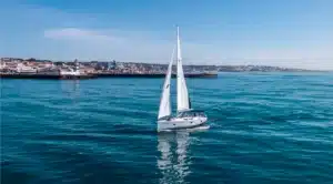 Yacht Katerina Wings with sails up, cruising near Cascais Marina on a serene day with clear blue sea and skyline, lighthouse guiding nearby.