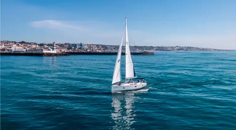 Yacht Katerina Wings with sails up, cruising near Cascais Marina on a serene day with clear blue sea and skyline, lighthouse guiding nearby.